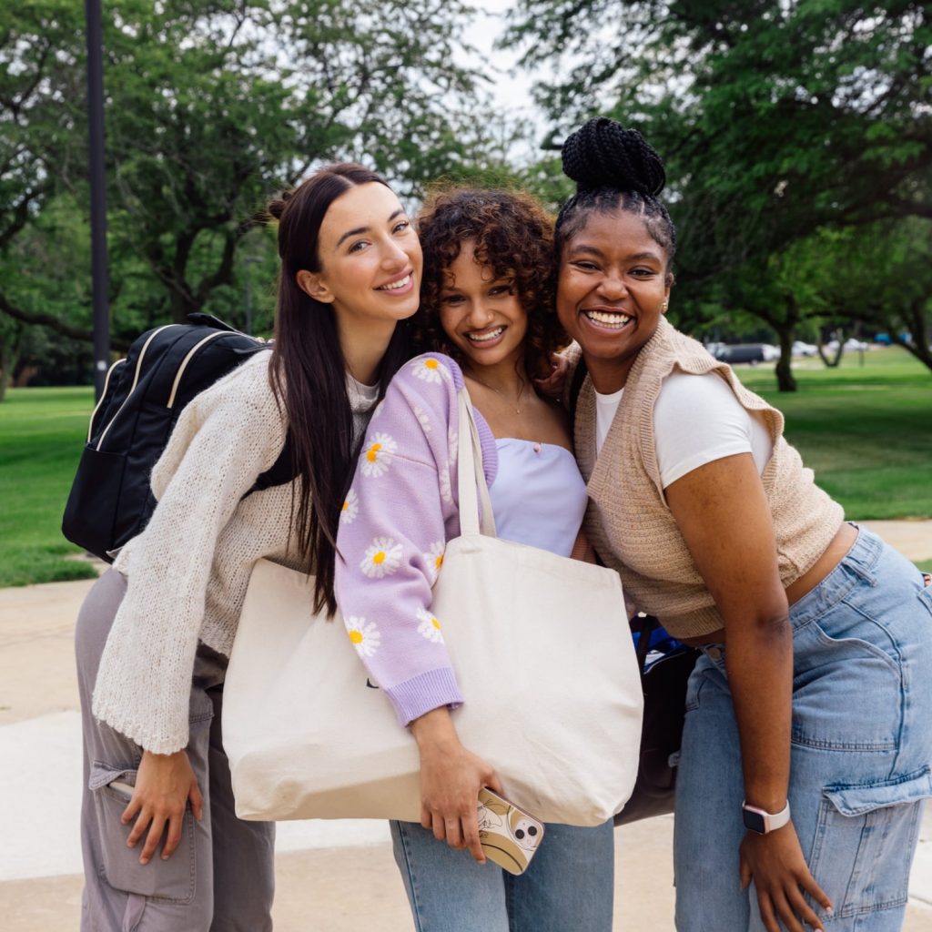 Three young women smile and lean together on an outdoor path with a grassy background: ​Left: Long dark hair, cream knit sweater, grey cargo pants, black backpack. ​Center: Curly brown hair, daisy-patterned purple cardigan, jeans, holding a large canvas tote bag. ​Right: Hair in a high bun, beige sweater vest over a white t-shirt, light-wash cargo jeans.