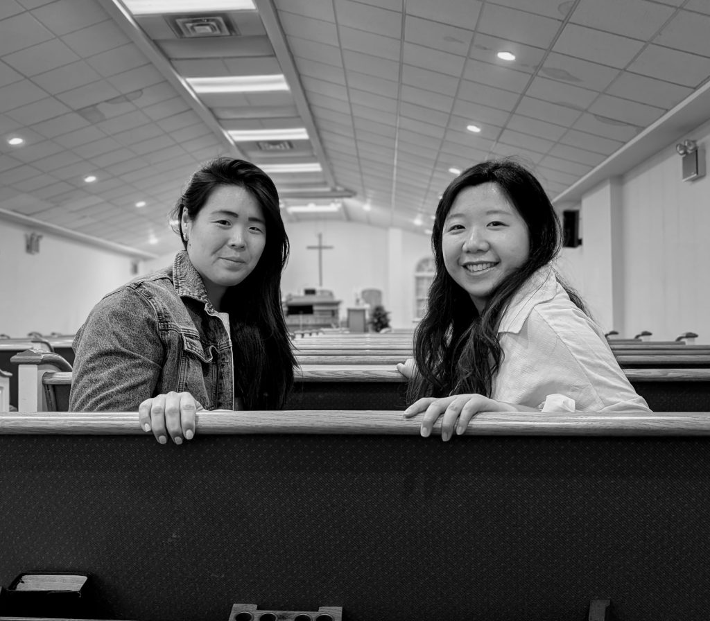 Two women are sitting in wooden church pews at Gracehouse Church community in Woodside, Queens. They are turning around to face the camera. The background shows rows of empty pews stretching toward an altar with a simple wooden cross mounted on the far wall under a vaulted ceiling.