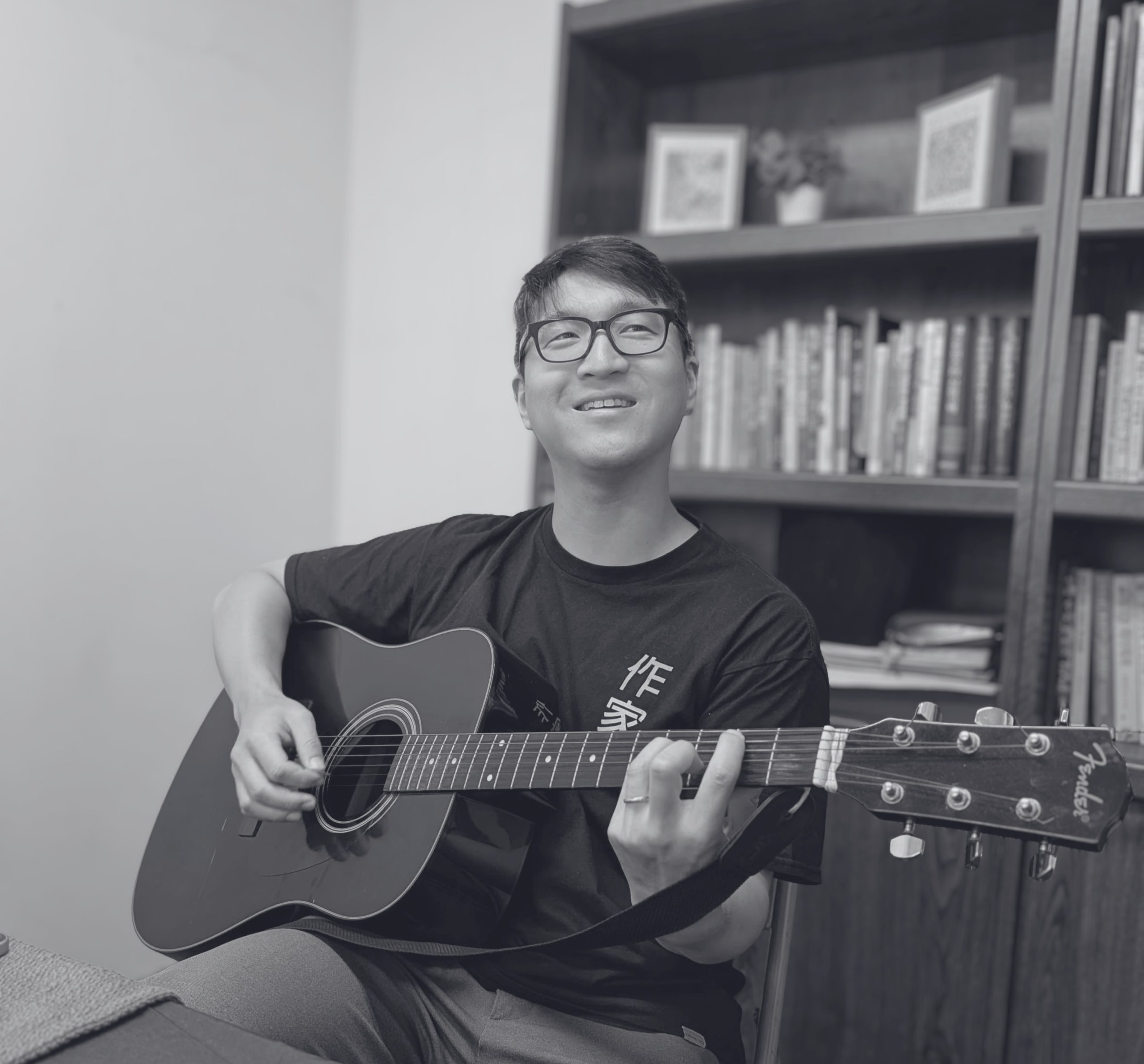 A man with short black hair and glasses is sitting down and smiling while playing a black Fender acoustic guitar. He is wearing a black t-shirt with yellow text and grey pants. A wooden bookshelf filled with books and a few small framed pictures sits in the background behind him